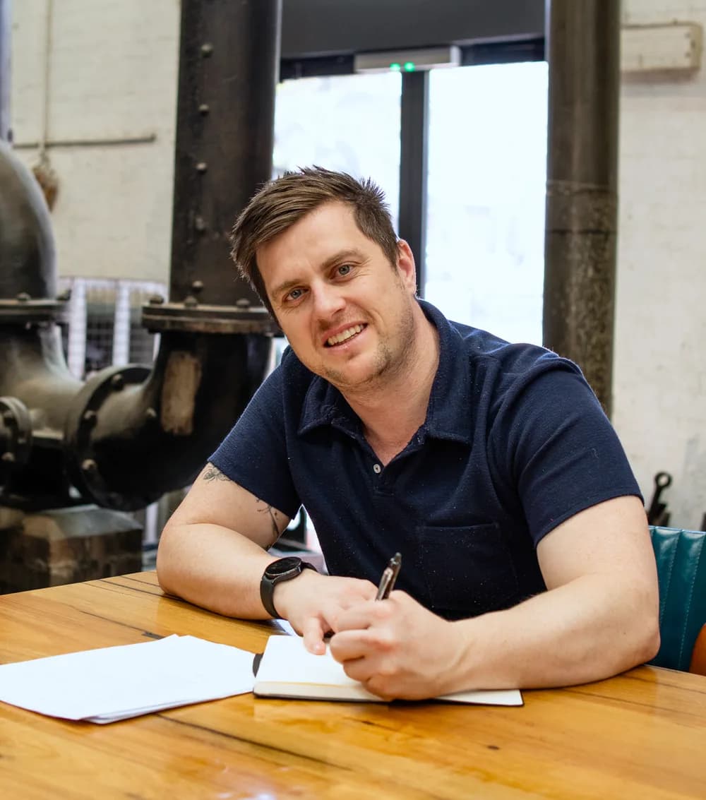 Brendan Smith at a timber table with a notebook and pen, papers beside him, smiling at the camera